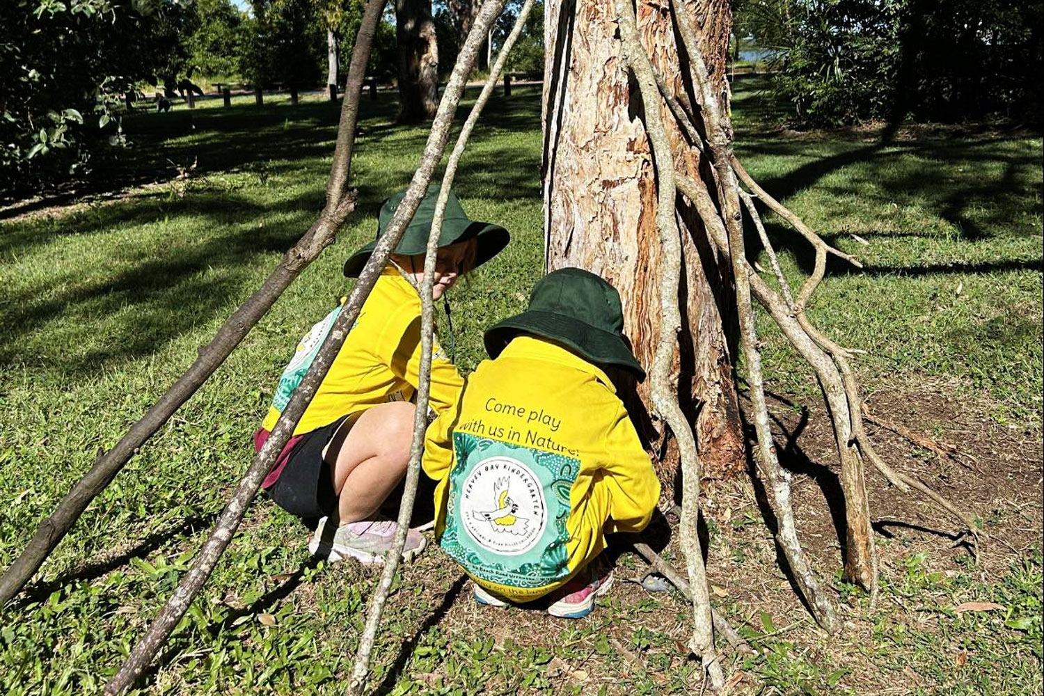 Two children playing outside next to a tree. They are building a cubby house with tree branches.