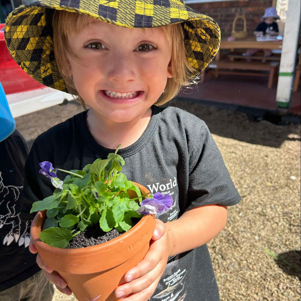 An overhead photograph of a young child drawing on a sheet of white paper at a wooden table. Next to her is an illustration showing the growth cycle of a tomato plant along with 3 pots of watercolours; yellow, green, and red. The child has used each of the watercolours on their paper and is now drawing lines with a black marker to recreate the tomato illustration.