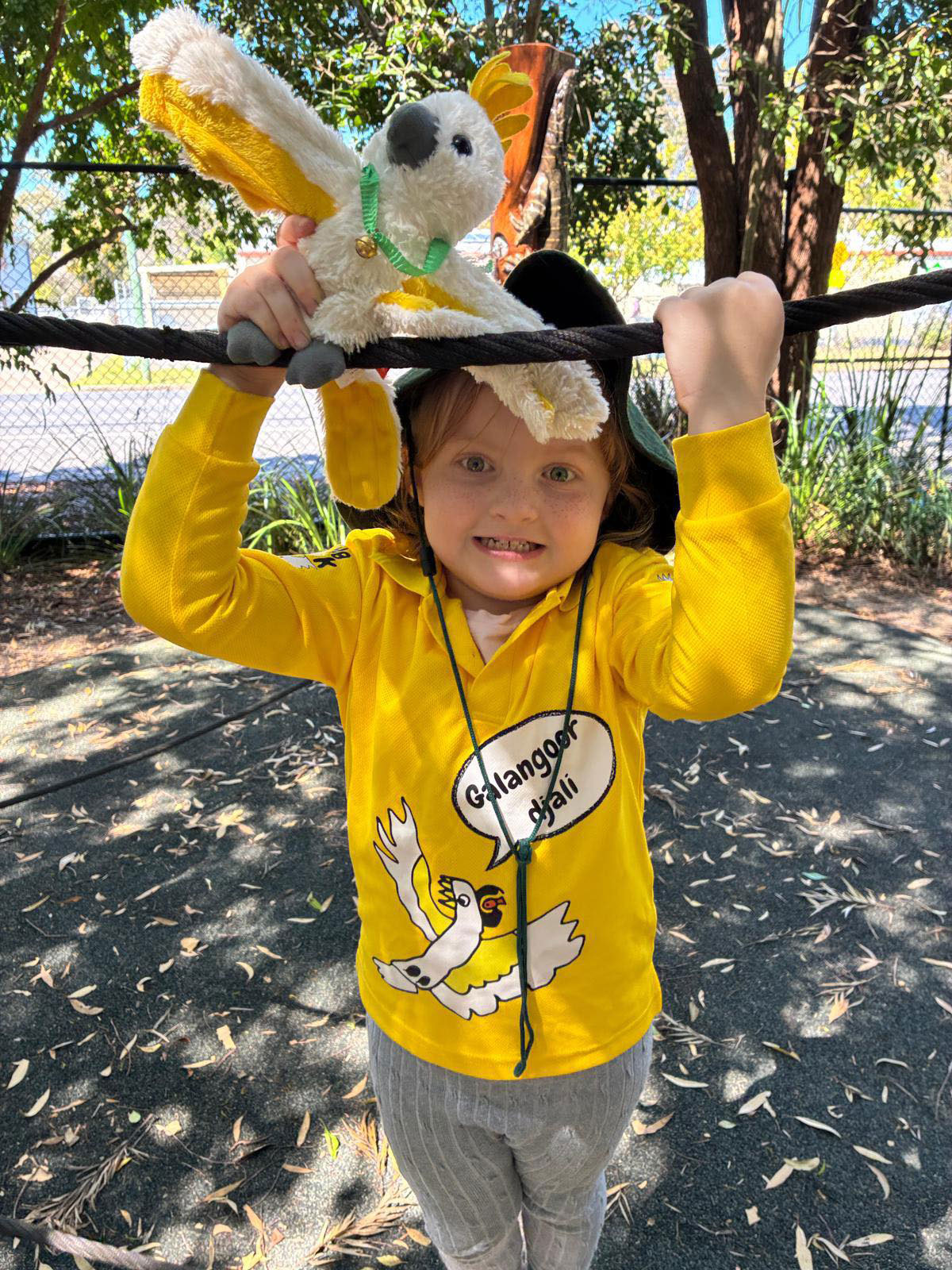 A young child in a long sleeve yellow shirt is playing with a stuffed Cockatoo toy outside